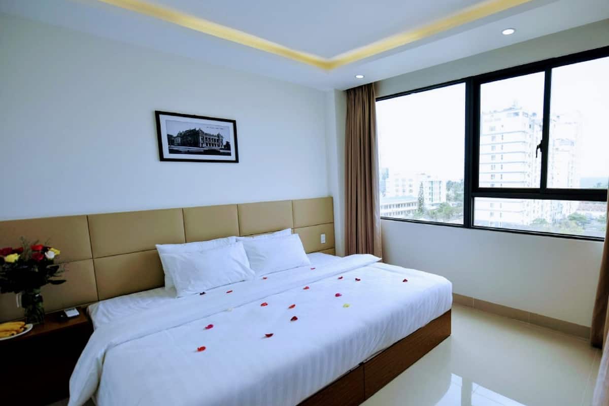 A hotel room with a large bed, a framed picture on the wall, and a view of buildings through the window.