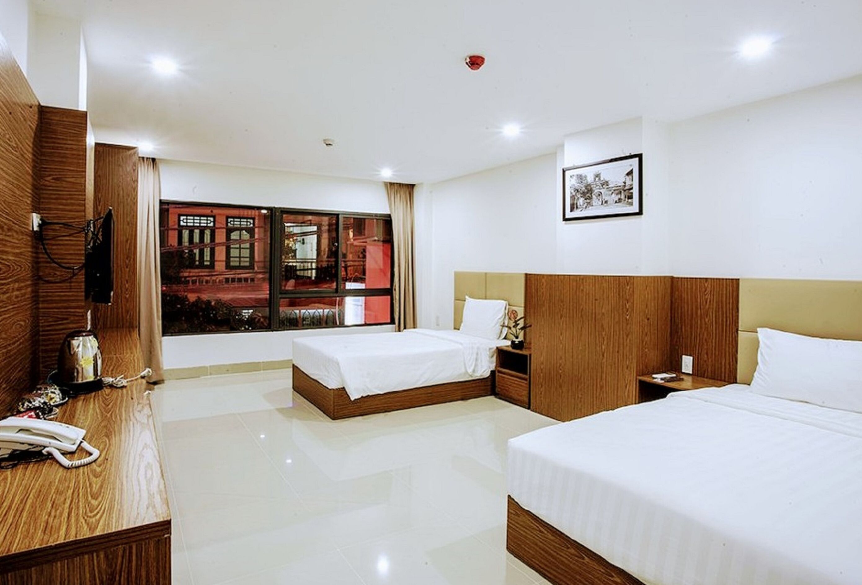 A hotel room with two beds, a wooden desk, a telephone, and a window with a view of buildings.
