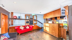 A lobby with a red sofa, a grey sofa, a wooden reception desk, and an ATM sign. - Snowy Valley - Hostel (East Jindabyne)