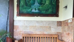 A wooden bench with a backrest, positioned in front of a stone wall with a framed painting of a waterfall. - Villa Sahabat (Gili Trawangan)