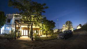 A hotel with a well-lit entrance, a covered outdoor area, and a dirt path leading to a parked SUV.