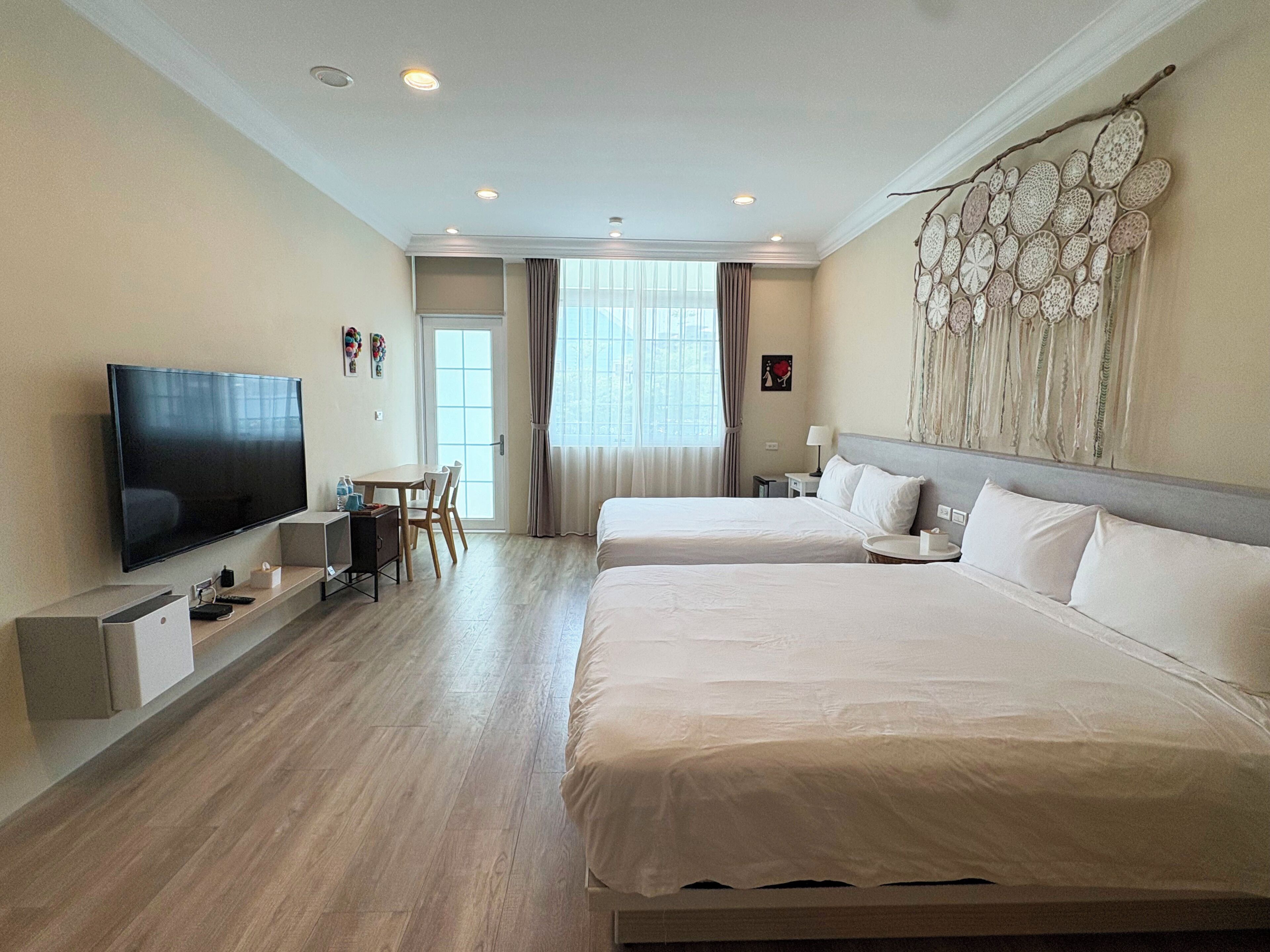 A hotel room with a flat-screen TV, two beds, a desk with a chair, and a large window with curtains.