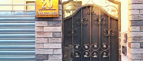 A brick building with a metal gate, a number sign '20 Macchiato', and a light fixture above the gate.