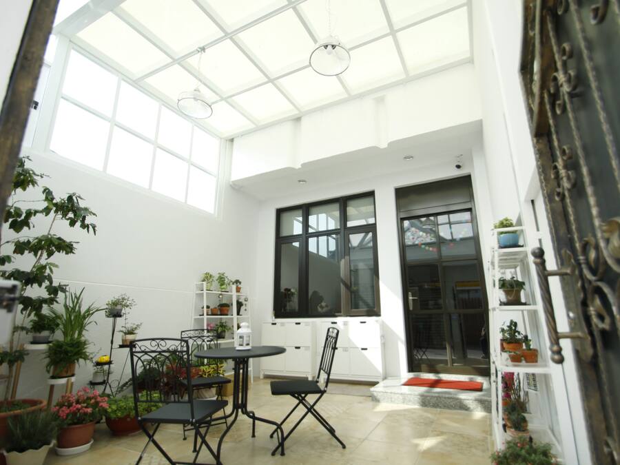 A bright, open-plan living area with a high ceiling and large windows, featuring a dining table and chairs, surrounded by potted plants.
