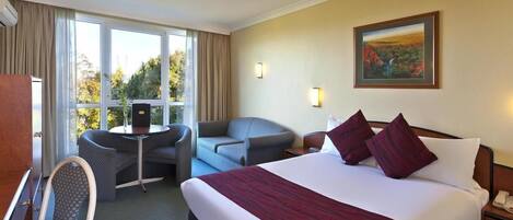 A hotel room with a large bed, two armchairs, a small table, and a view of the outdoors through large windows.