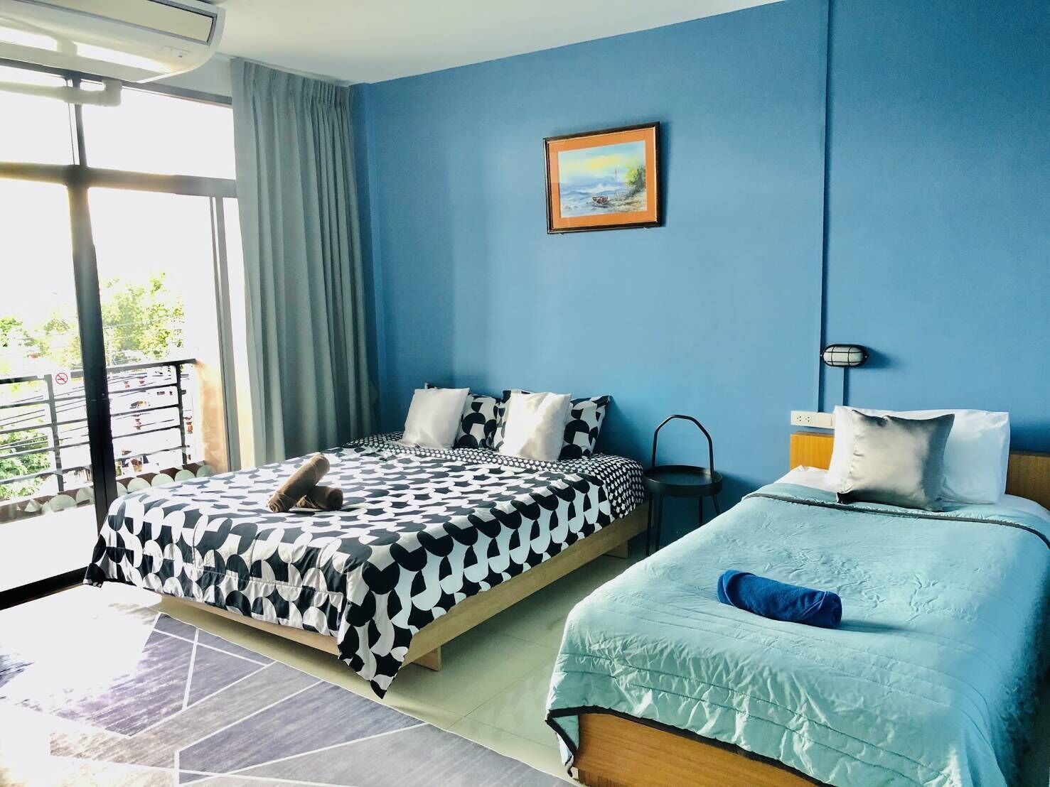 A hotel room with two beds, a large window with curtains, a painting on the wall, and a balcony with a railing.