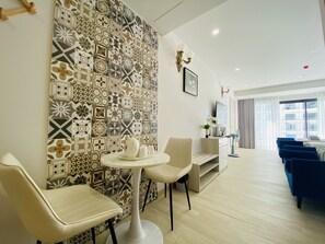 Quarto de hotel moderno com parede estampada, mesa redonda com cadeiras, televisão e vista da cidade por grandes janelas.