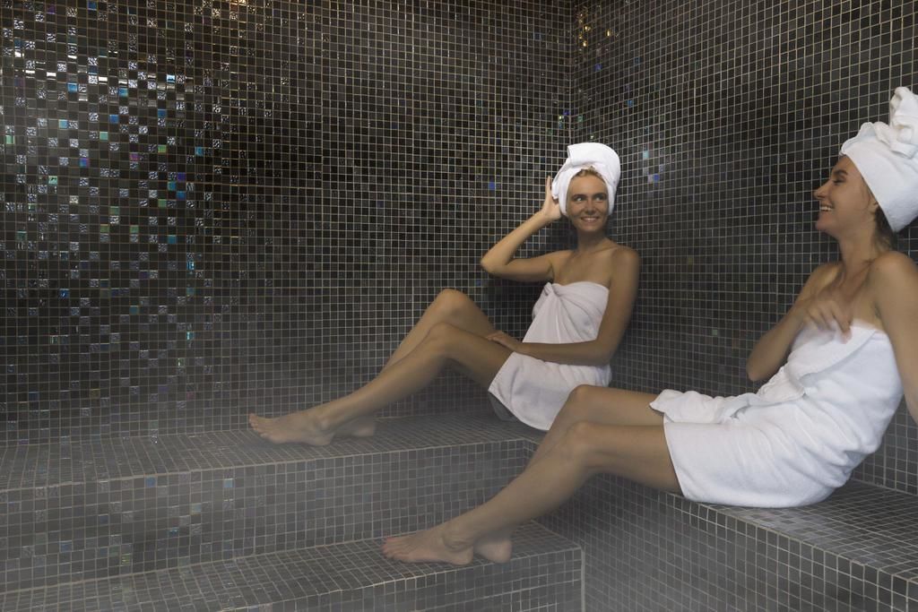 Two individuals seated in a tiled room with a mosaic wall.