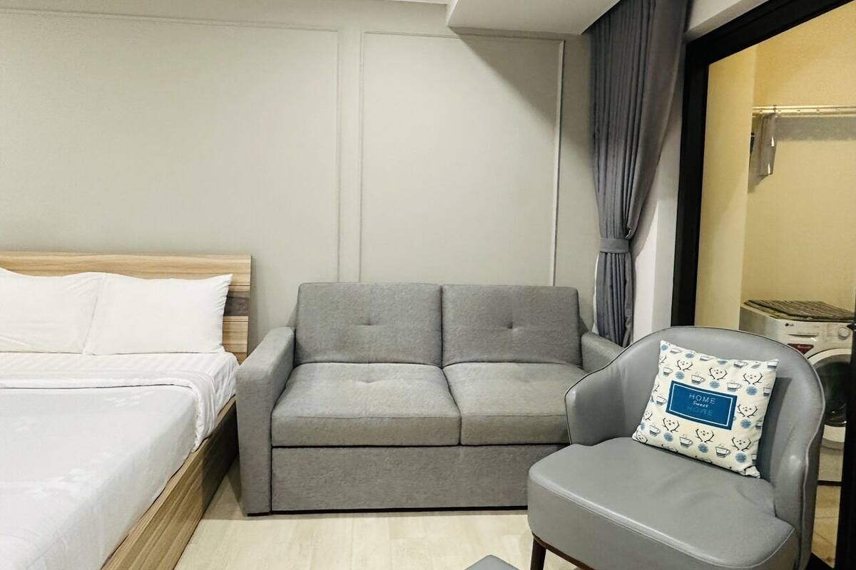 Una habitación de hotel con una cama, un sofá gris, un sillón gris y una mesita de noche con una lámpara.