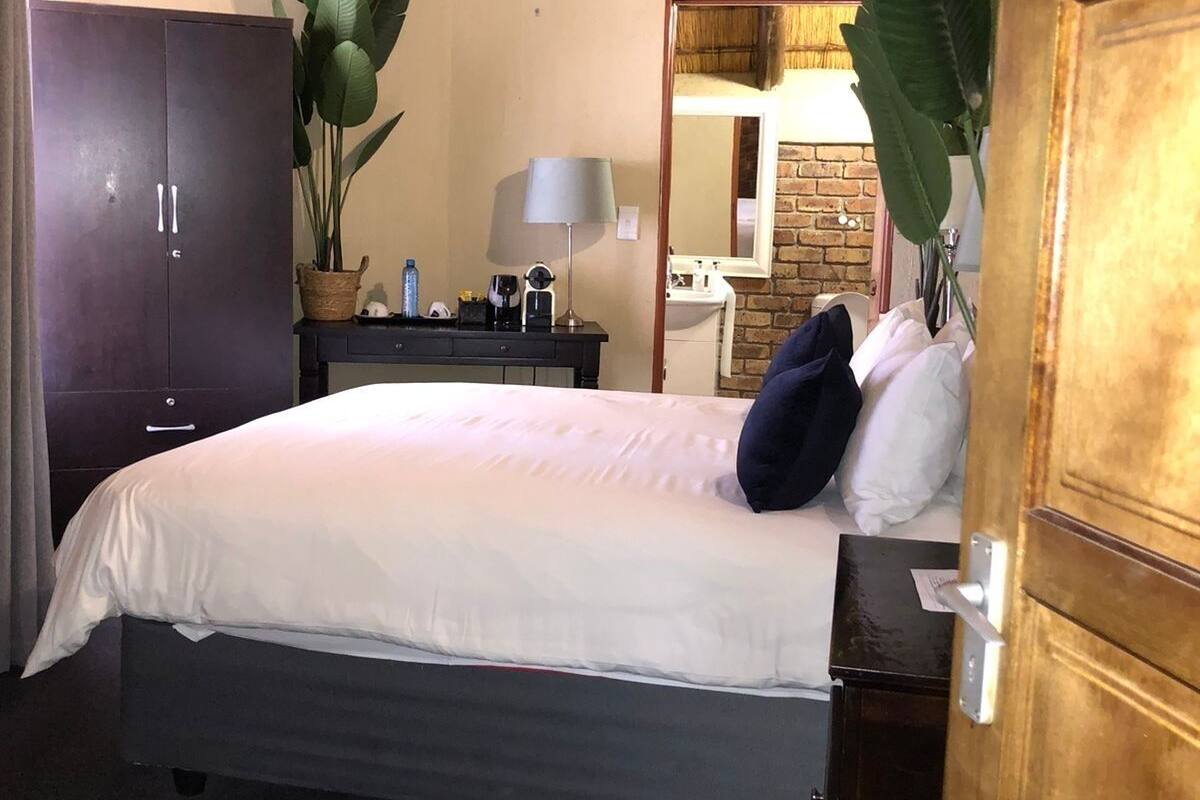 A hotel room with a bed, a wardrobe, a desk with a lamp, a mirror, and a potted plant.