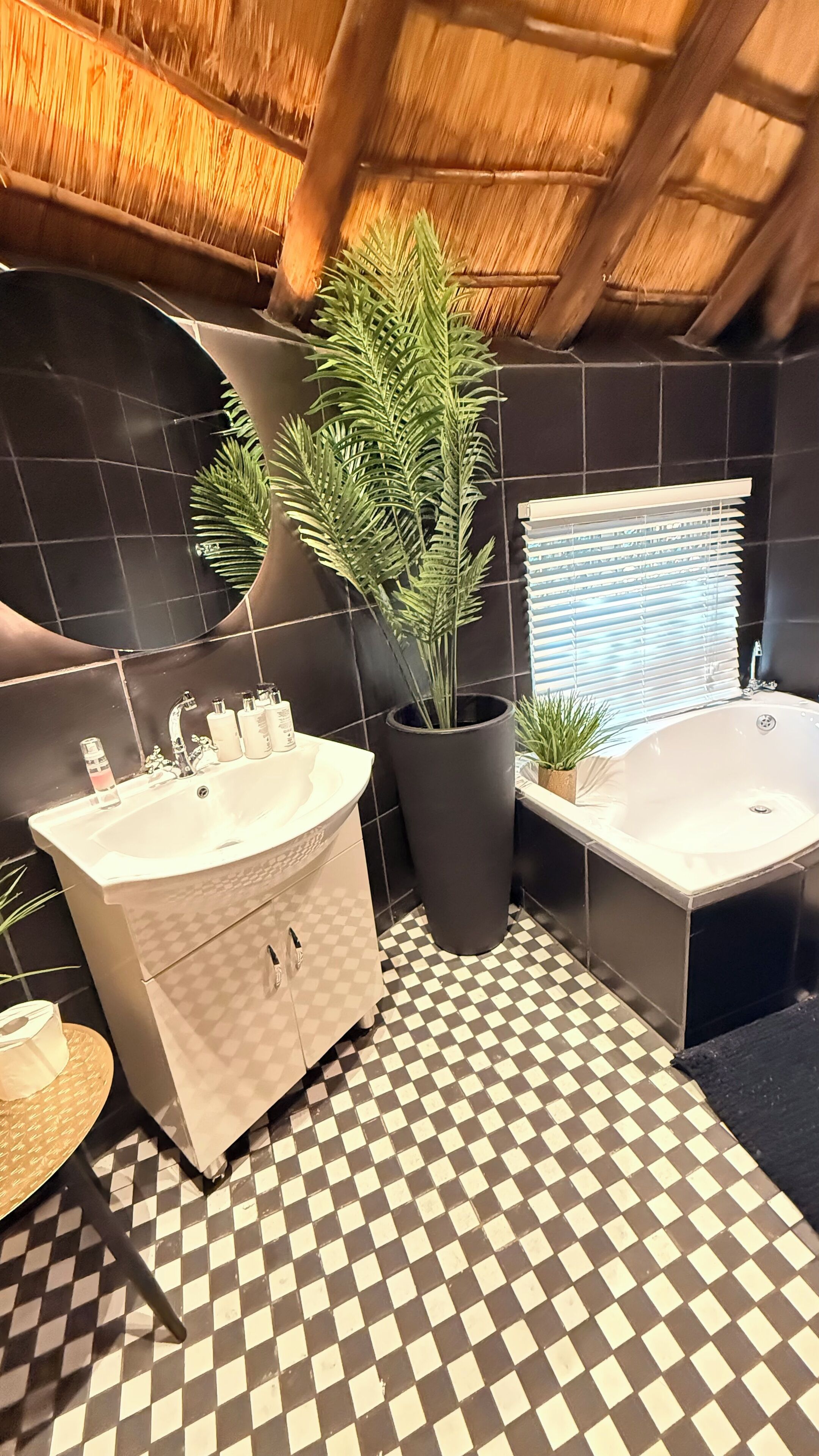 A bathroom with a checkered floor, a large potted plant, a bathtub, and a vanity with a mirror.
