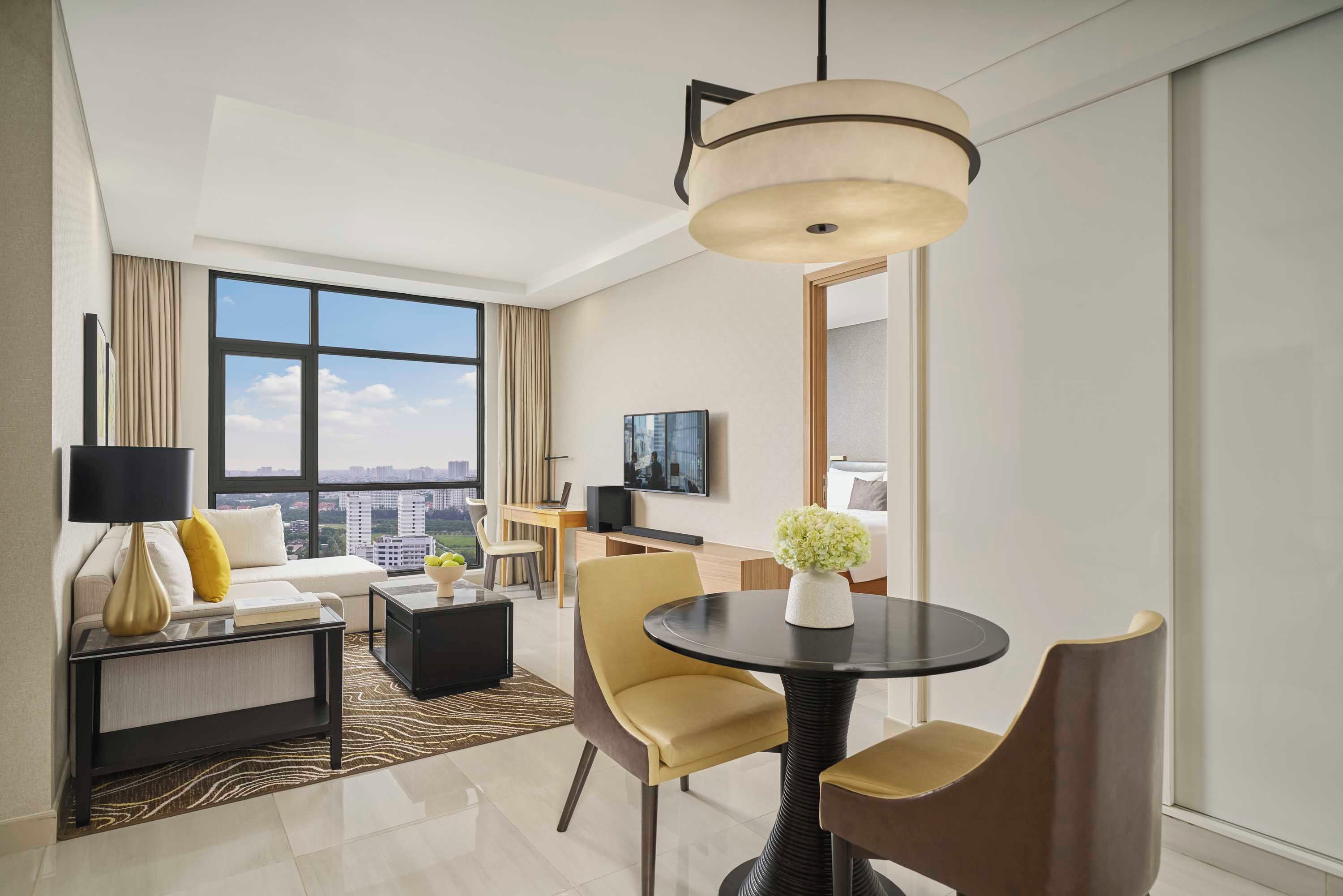 A modern hotel room with a large window, a dining area, a sofa, a TV, and a decorative vase.