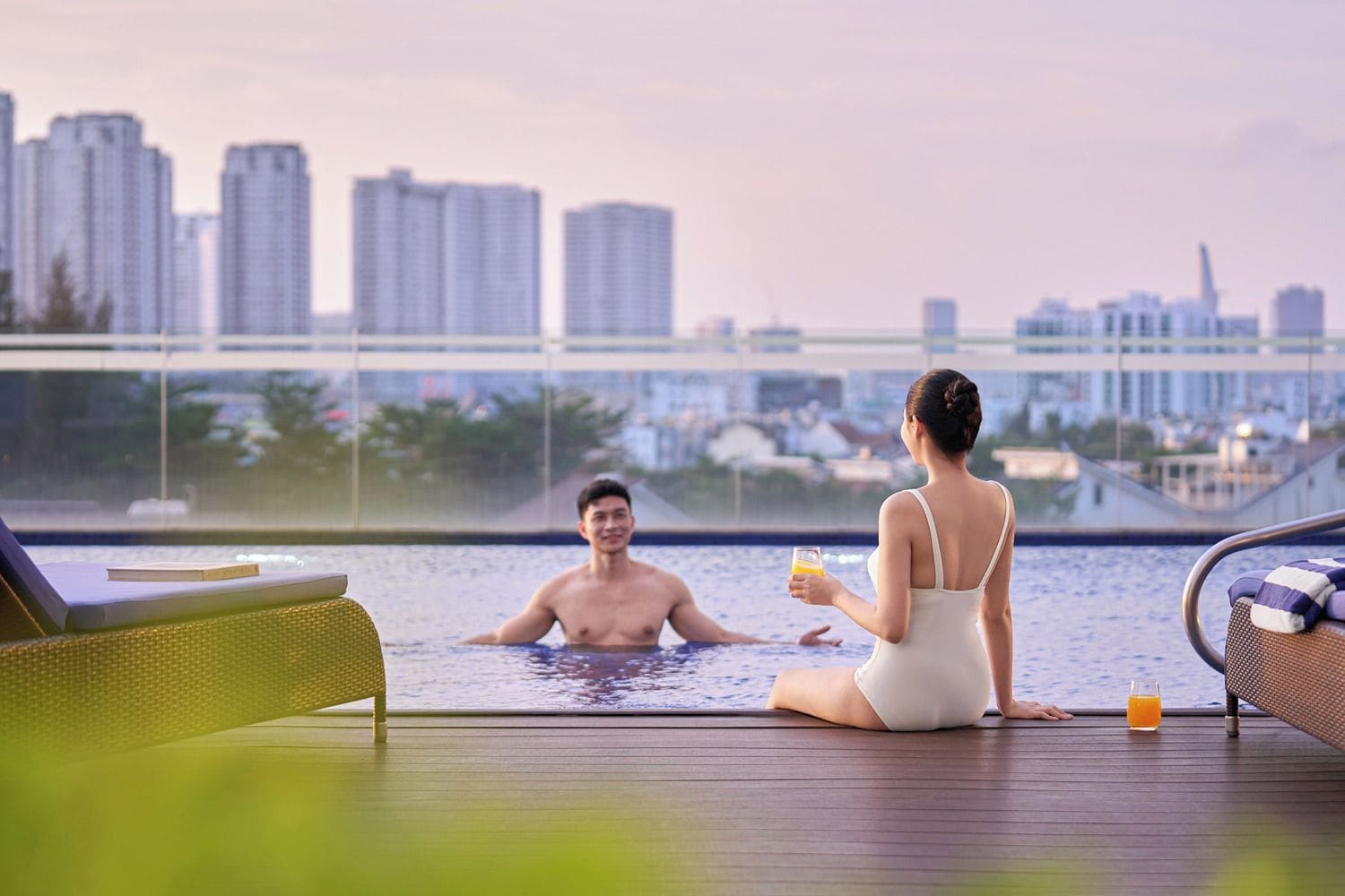 A couple relaxing by a pool with a cityscape view, a lounge chair, and a glass of drink on the deck.