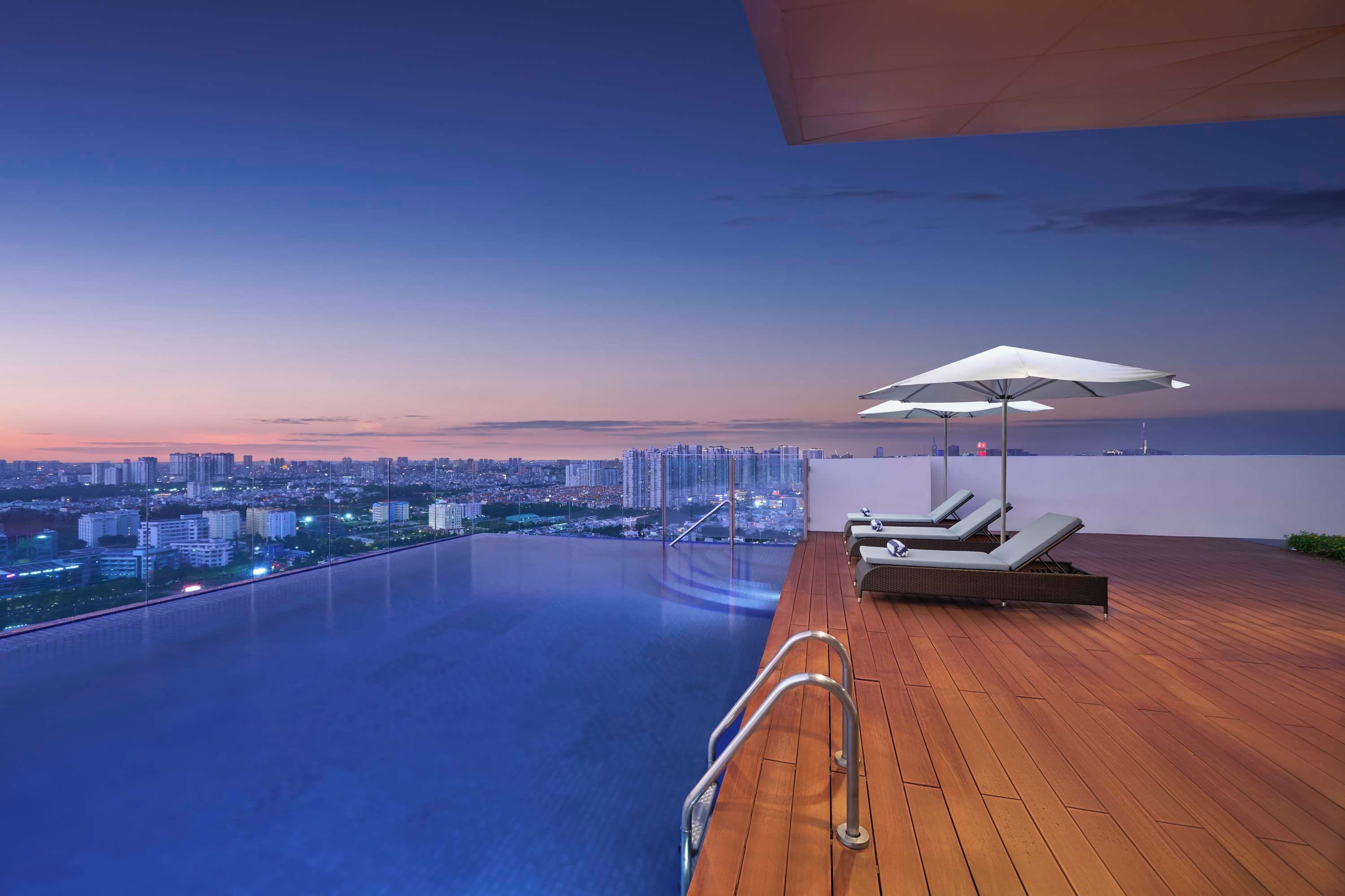Una piscina infinita con muelle de madera y sillones de descanso, con vista a un paisaje urbano al atardecer.