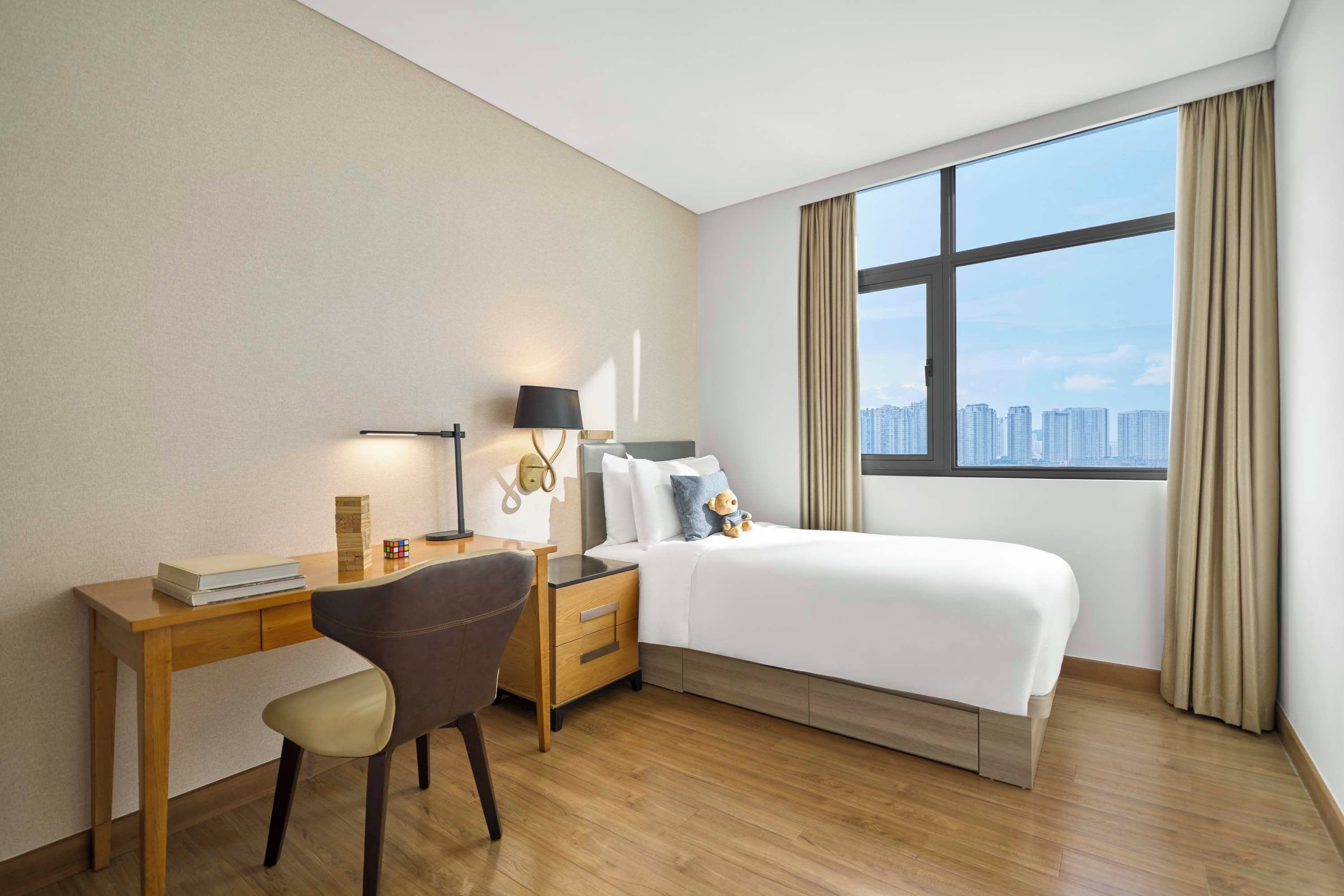 A hotel room with a bed, a desk, a chair, a lamp, and a view of the city through a large window.