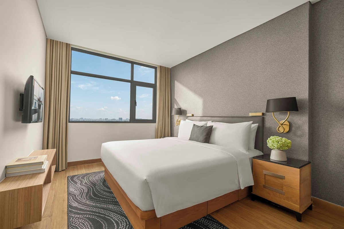 Une chambre d’hôtel avec un grand lit, une table de chevet avec une lampe, un téléviseur fixé au mur et une vue sur le paysage urbain par la fenêtre.