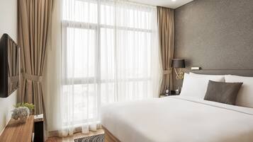 A modern hotel room with a large bed, a flat-screen TV mounted on the wall, a bedside table with a lamp, and sheer curtains on the window.