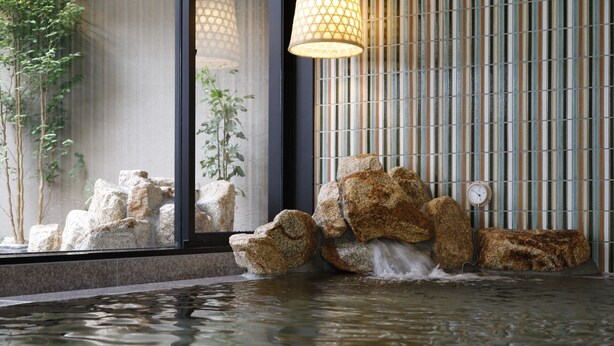 Un élément décoratif aquatique composé de rochers et d’une horloge, placé devant un mur à rayures et une fenêtre ornée d’une plante.