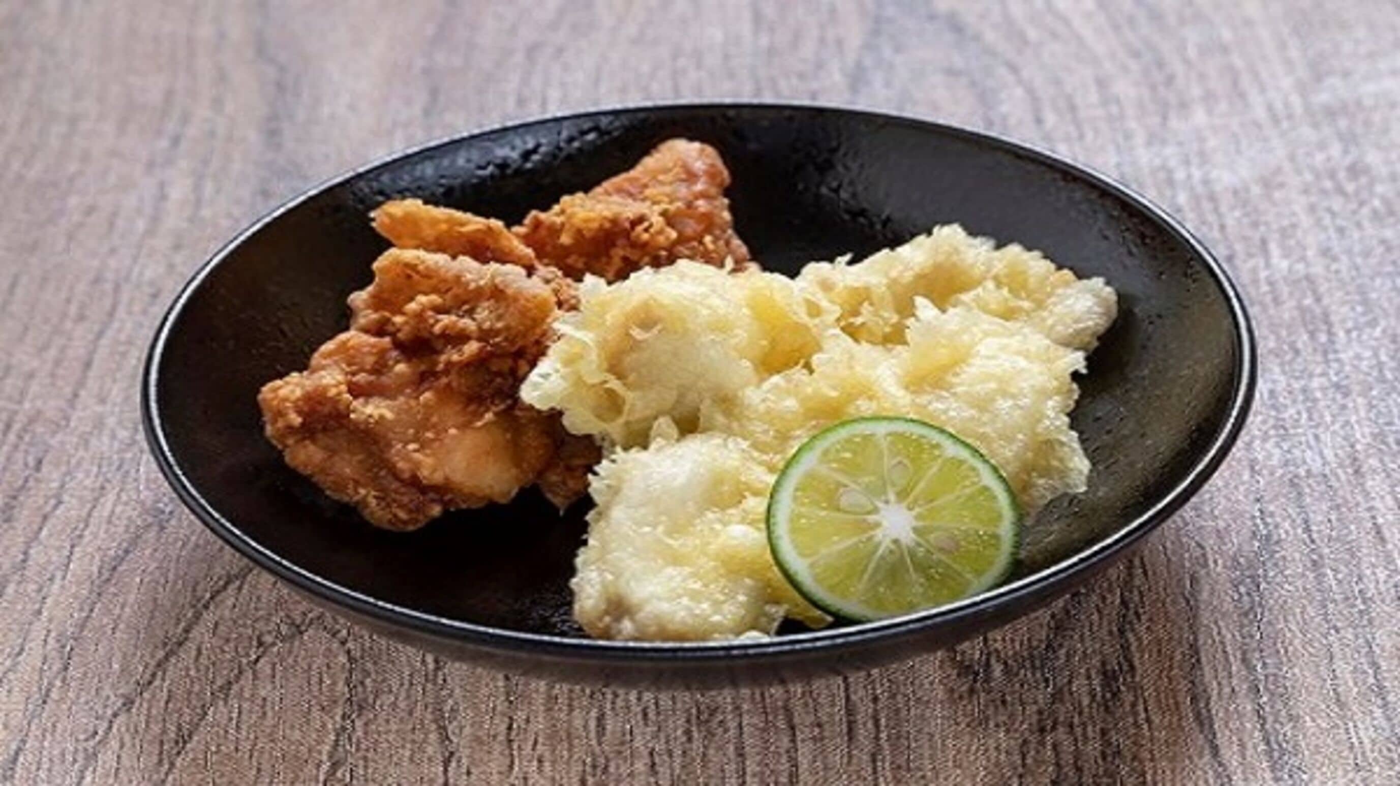 Een zwart bord met gefrituurde kip, puree en een stukje limoen op een houten tafel.