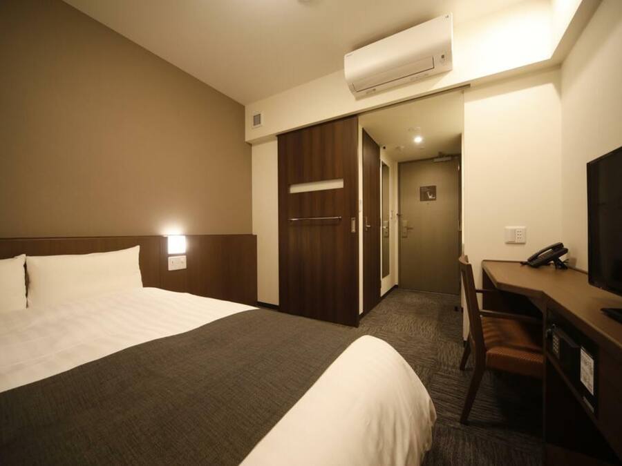 Una habitación de hotel con una cama grande, un escritorio con silla, un televisor y un aire acondicionado.
