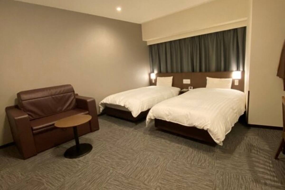 Ein Hotelzimmer mit zwei Betten, einem braunen Ledersofa, einem kleinen runden Tisch und einer Deckenleuchte.