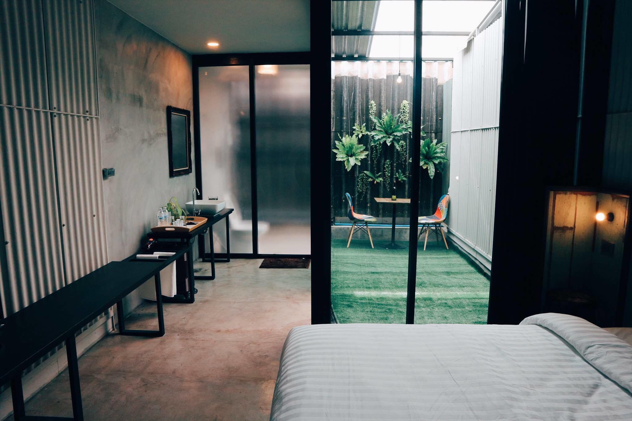 A modern bedroom with a large bed, a desk, and a chair. There is a sliding glass door leading to a small outdoor area with plants.