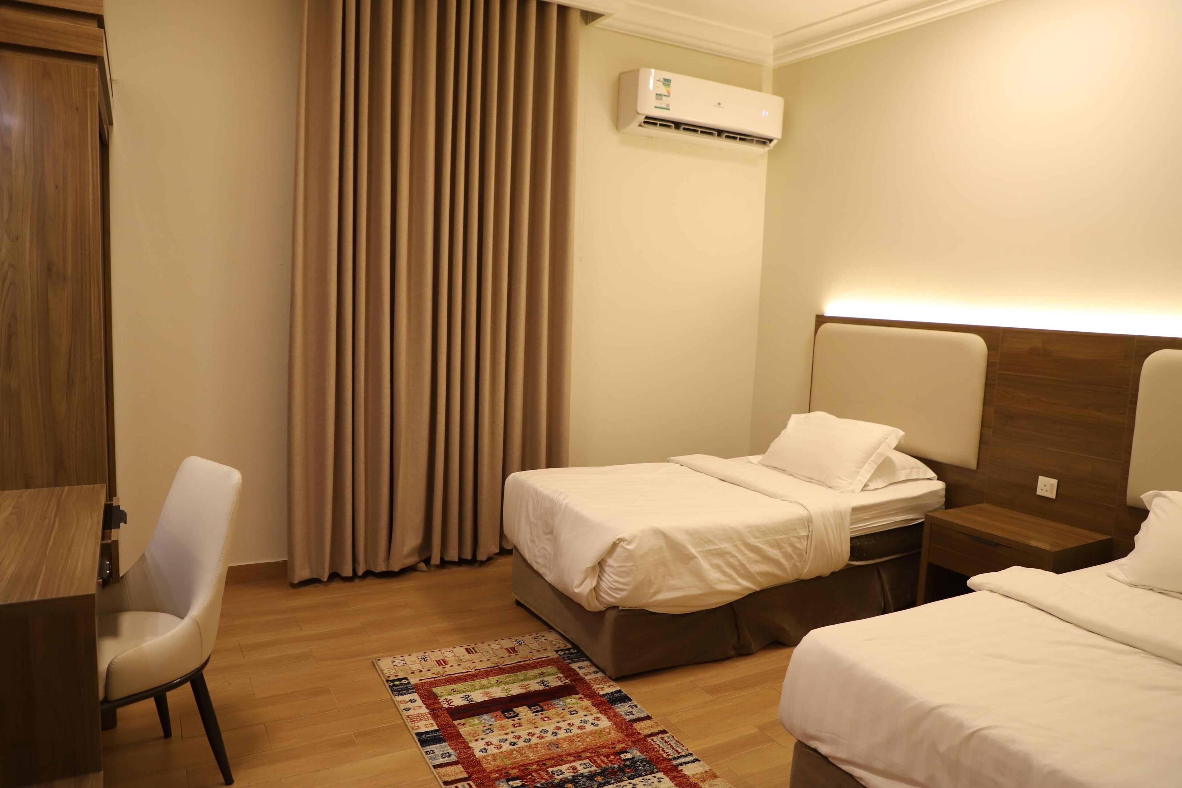 A hotel room with two beds, a desk, a chair, an air conditioning unit, and a patterned rug.
