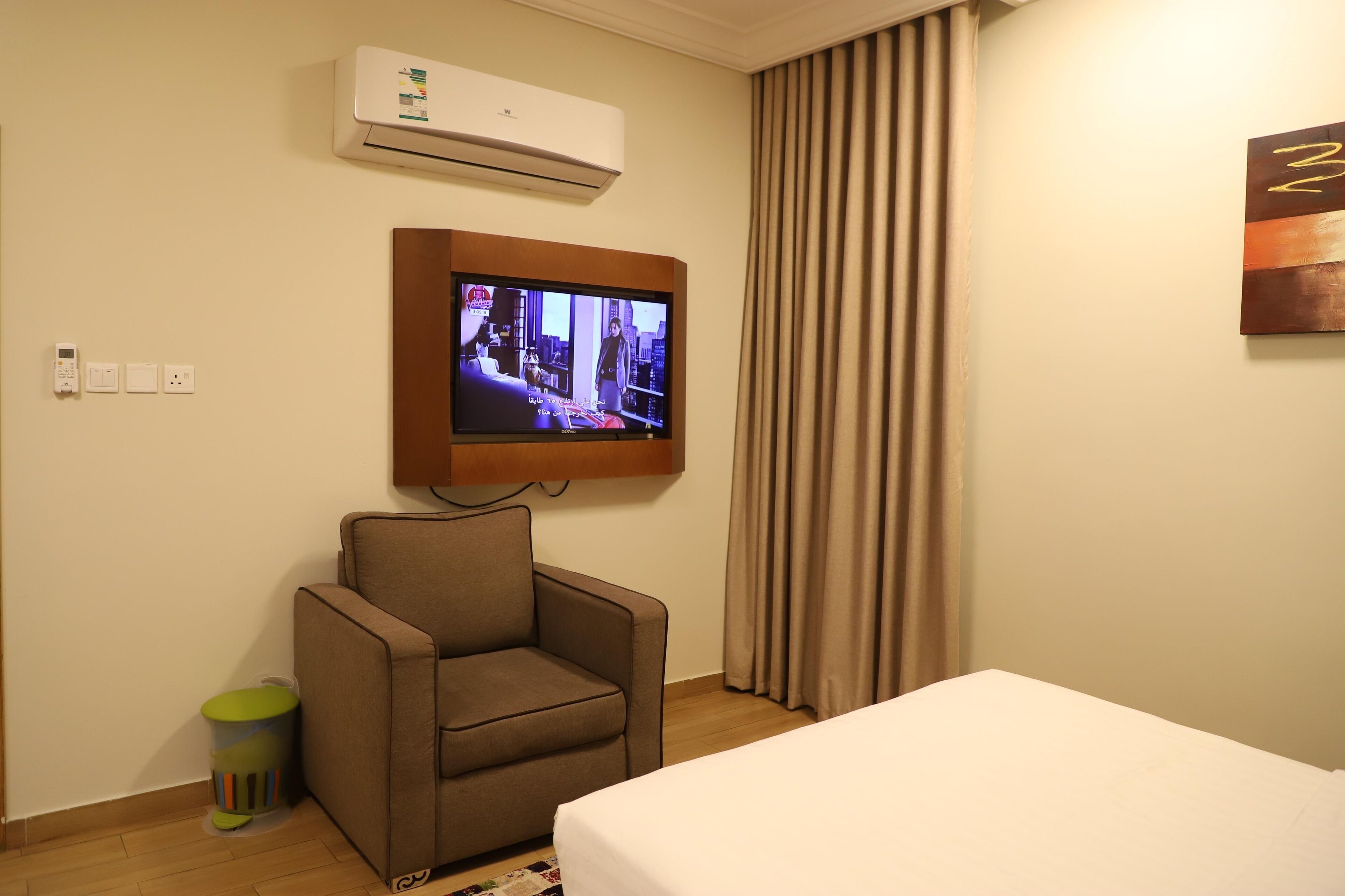 A hotel room with a bed, a brown armchair, a television mounted on the wall, an air conditioning unit, and a curtain.
