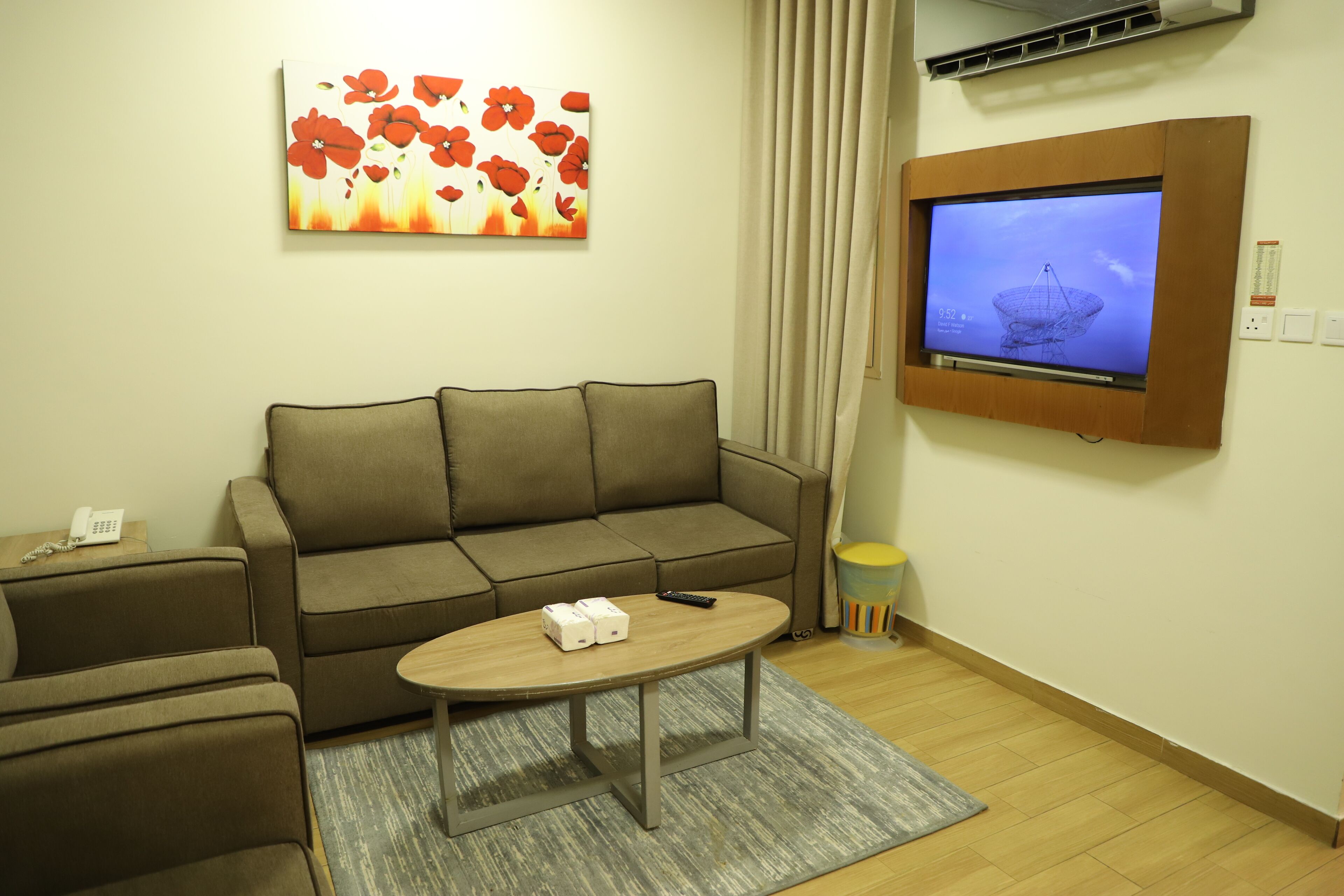 A living room with a sofa, a coffee table, a flat-screen TV mounted on the wall, a painting, and a small trash bin.