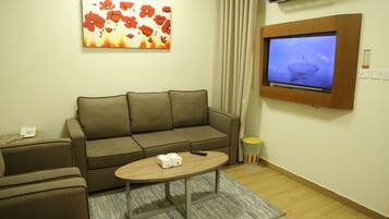 A living room with a sofa, a coffee table, a flat-screen TV mounted on the wall, a painting, and a small trash bin.