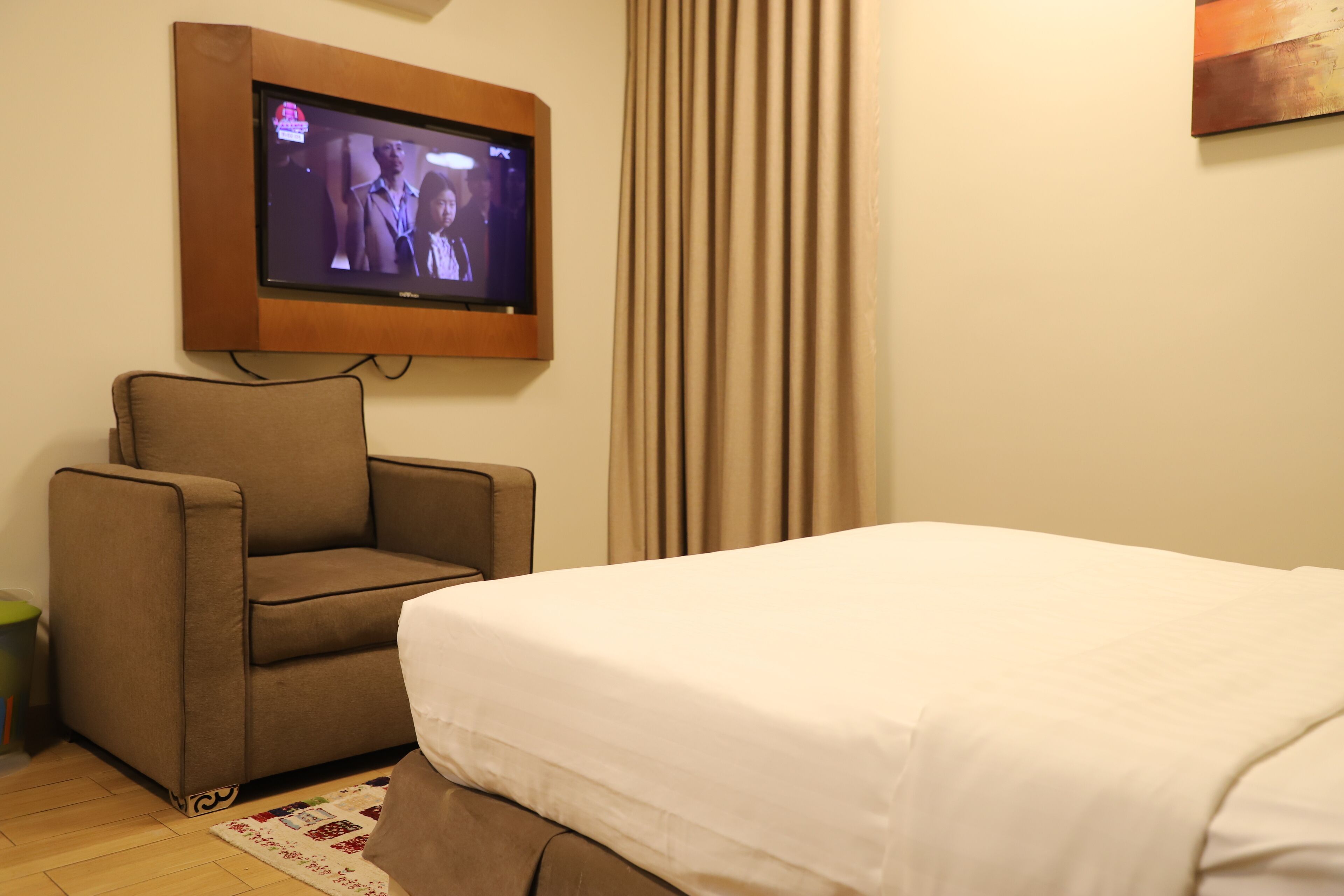 Una habitación de hotel con una cama, un sillón marrón, un televisor montado en la pared y una cortina.
