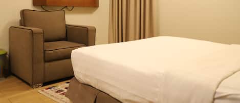 Una habitación de hotel con una cama, un sillón marrón, un televisor montado en la pared y una cortina.