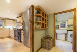 Une cuisine équipée d’un four encastré, d’un réfrigérateur, d’étagères en bois où sont rangés de la vaisselle, et d’un lave-linge dans la pièce adjacente.