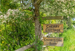 A wooden signpost with two signs, one reading 'Cherry Cottage' and the other 'Cherrystone Barn,' surrounded by lush greenery.