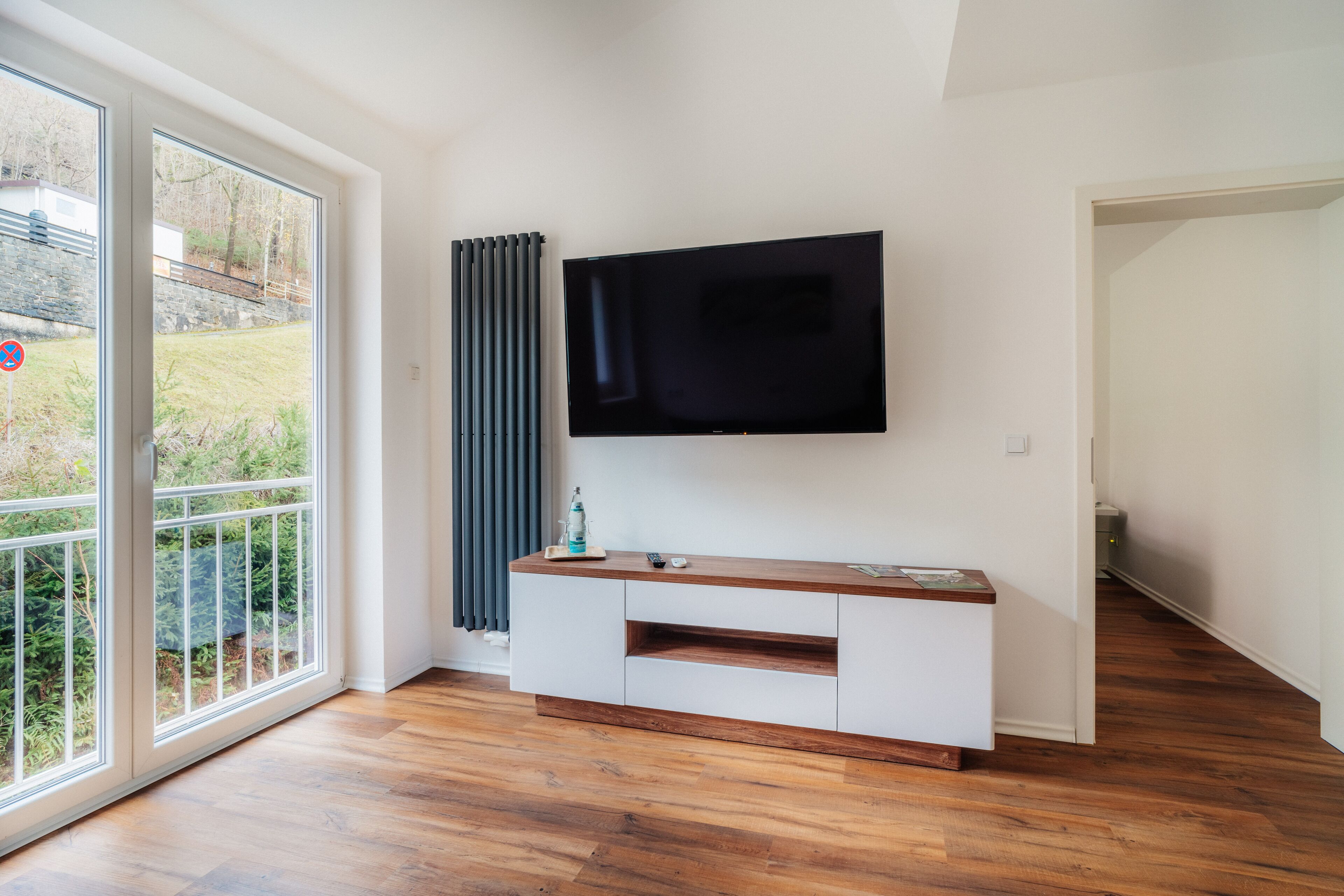 Ein modernes Wohnzimmer mit einem großen Flachbildfernseher an der Wand, einer weißen Unterhaltungseinheit mit Holzplatte und einer Glastür, die auf einen Balkon mit Blick auf Grünflächen und eine Steinmauer führt.