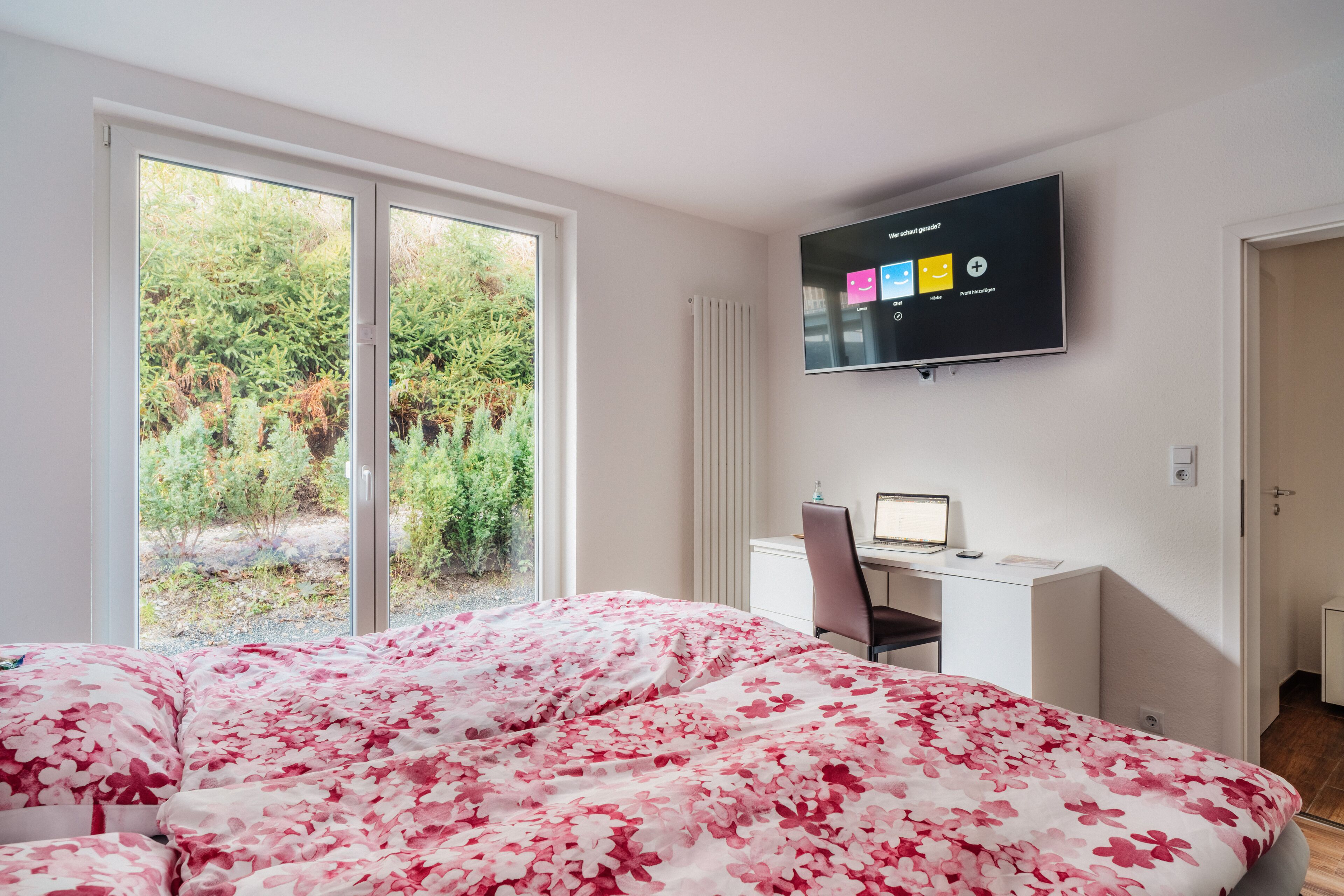 A bedroom with a bed, a desk, a chair, a TV, and a window with a view of greenery.