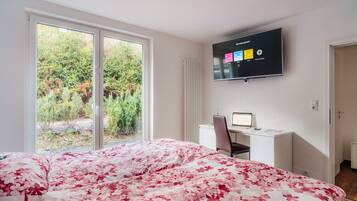 Un dormitorio con cama, escritorio, silla, televisor y ventana con vista a vegetación.
