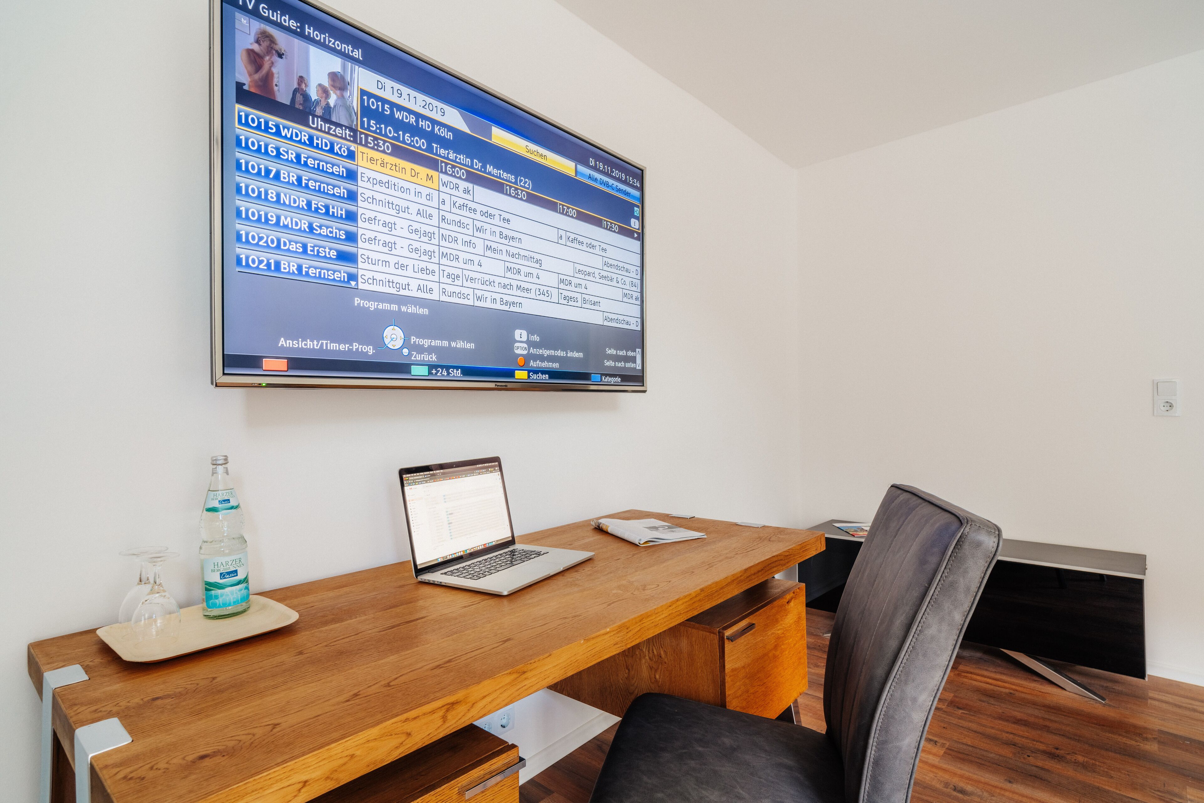 A modern office space with a wooden desk, a laptop, and a large TV screen displaying a TV guide.