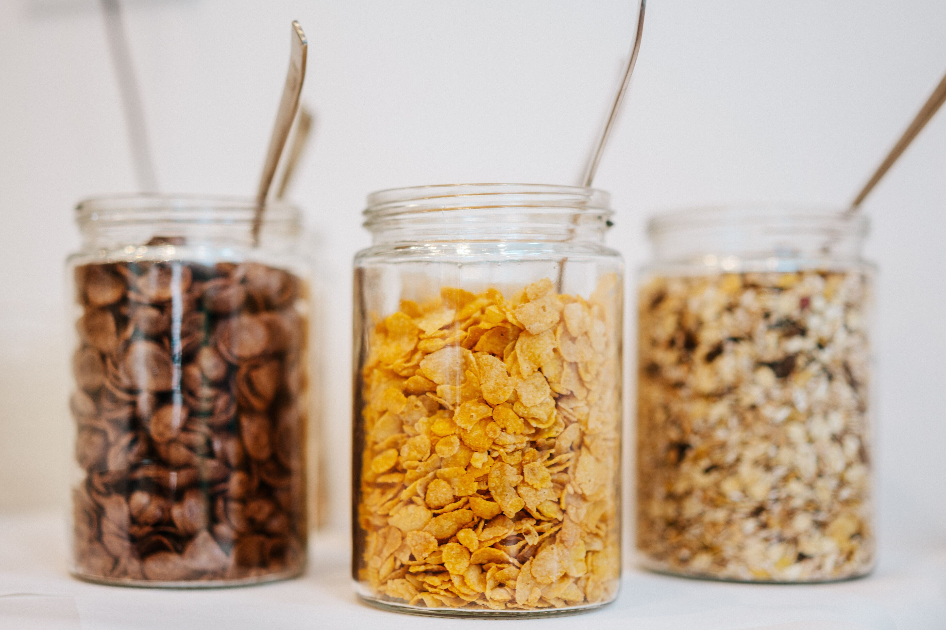 Three glass jars filled with different types of cereal, each with a spoon.