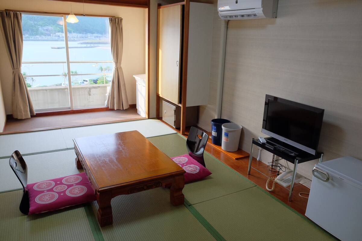 A room with tatami mats, a wooden table, a television, a refrigerator, and a view of a body of water.