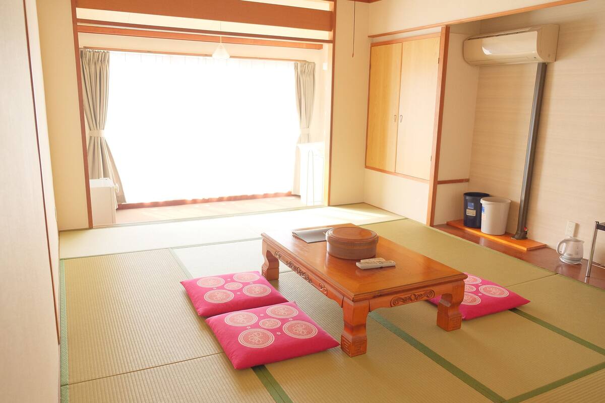 A traditional Japanese room with tatami flooring, a wooden table with cushions, a window with curtains, and a radiator.