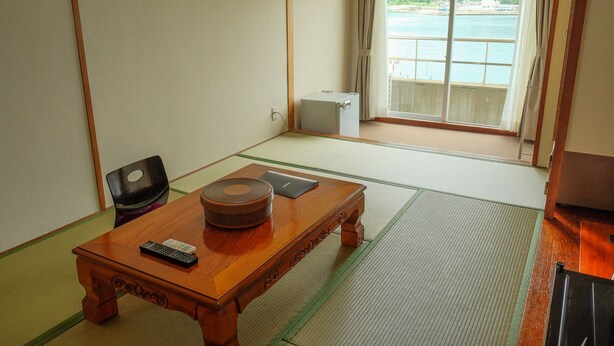 A traditional Japanese room with tatami flooring, a wooden table with a remote control and a book, and a view of a body of water through a sliding door. - Sunset Susami (Susami)