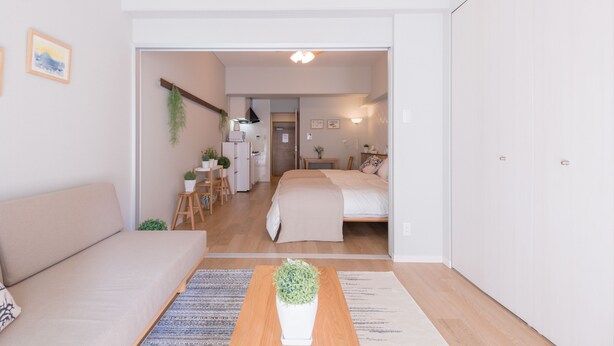 Una habitación moderna y bien iluminada, con un sofá, una mesa de centro de madera con una planta, una cama con colcha color beige y un escritorio con silla y una planta colgante.