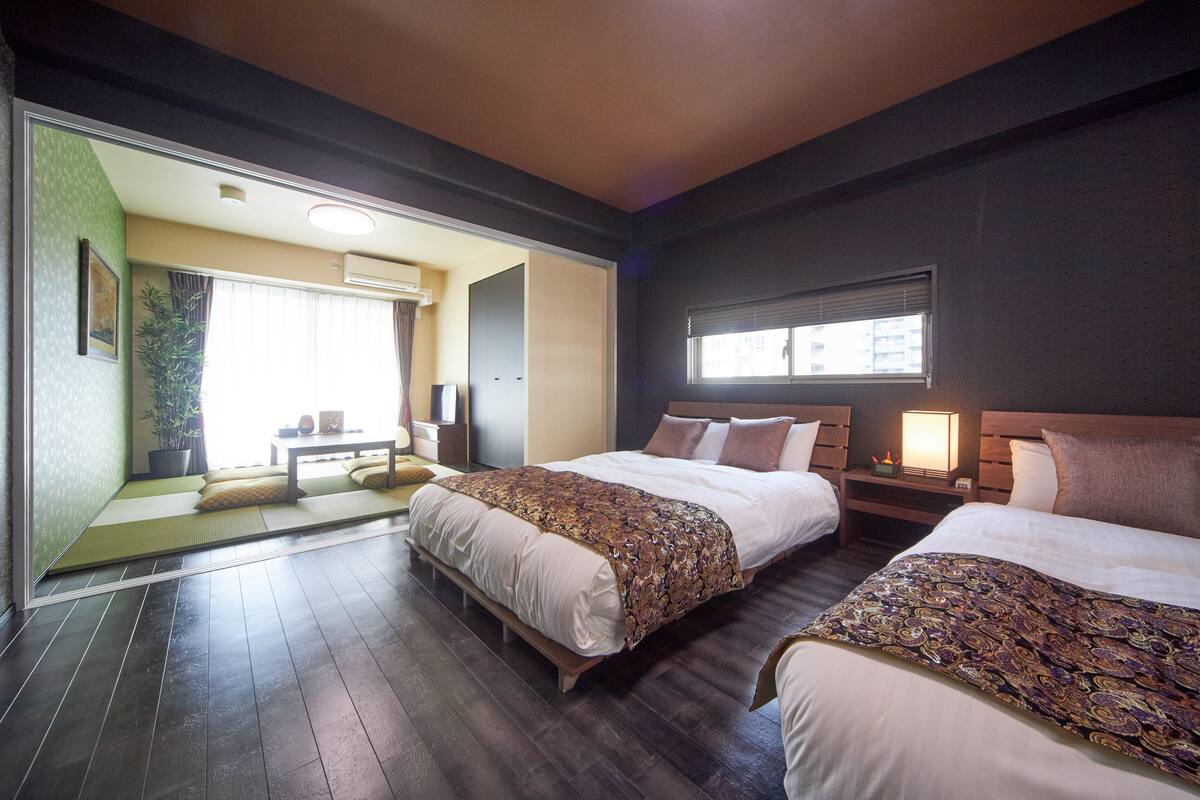 A hotel room with two beds, a sitting area with a tatami mat, a potted plant, and a window with blinds.