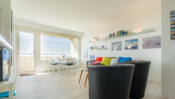 Una sala moderna con un sofá negro, un comedor con sillas blancas y un amplio ventanal con vistas al cielo.