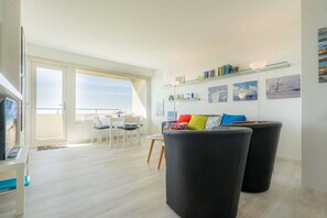 Una sala moderna con un sofá negro, un comedor con sillas blancas y un amplio ventanal con vistas al cielo.