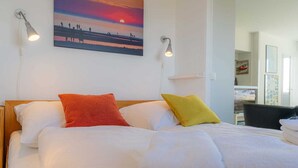 Una cama bien hecha con sábanas blancas y dos almohadas, una roja y otra amarilla, en una habitación con un cuadro de un atardecer y dos luces empotradas en la pared.