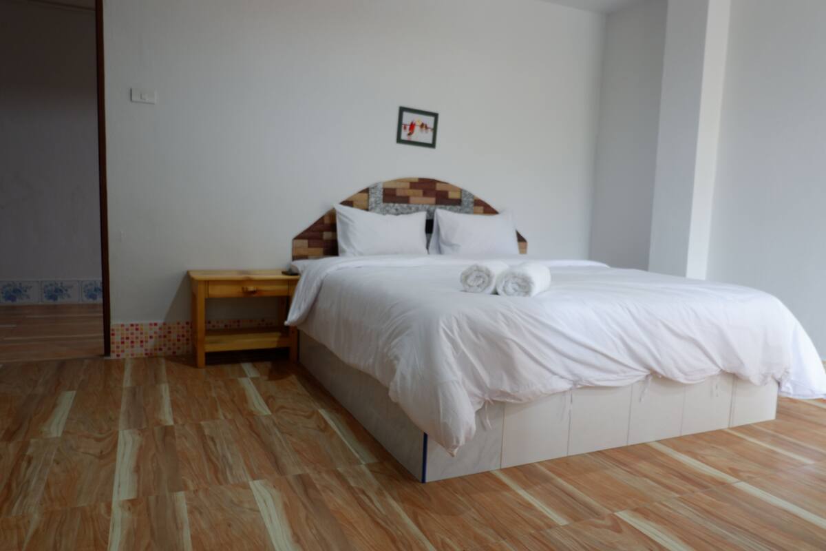 A bedroom with a wooden floor, a bed with white bedding, a nightstand, and a wall-mounted picture.
