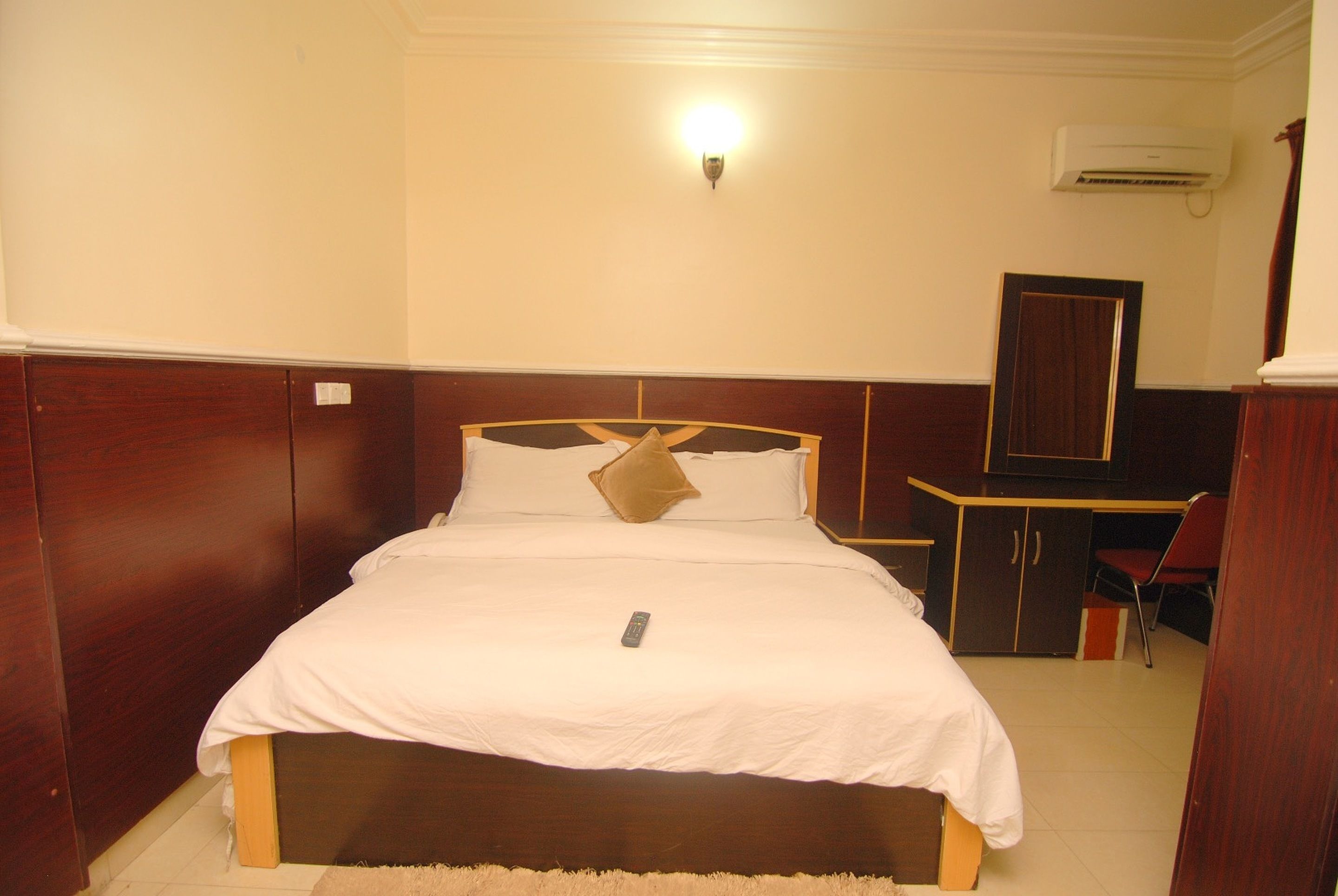 A neatly made bed with a remote control on it, a desk with a chair, and an air conditioning unit on the wall.
