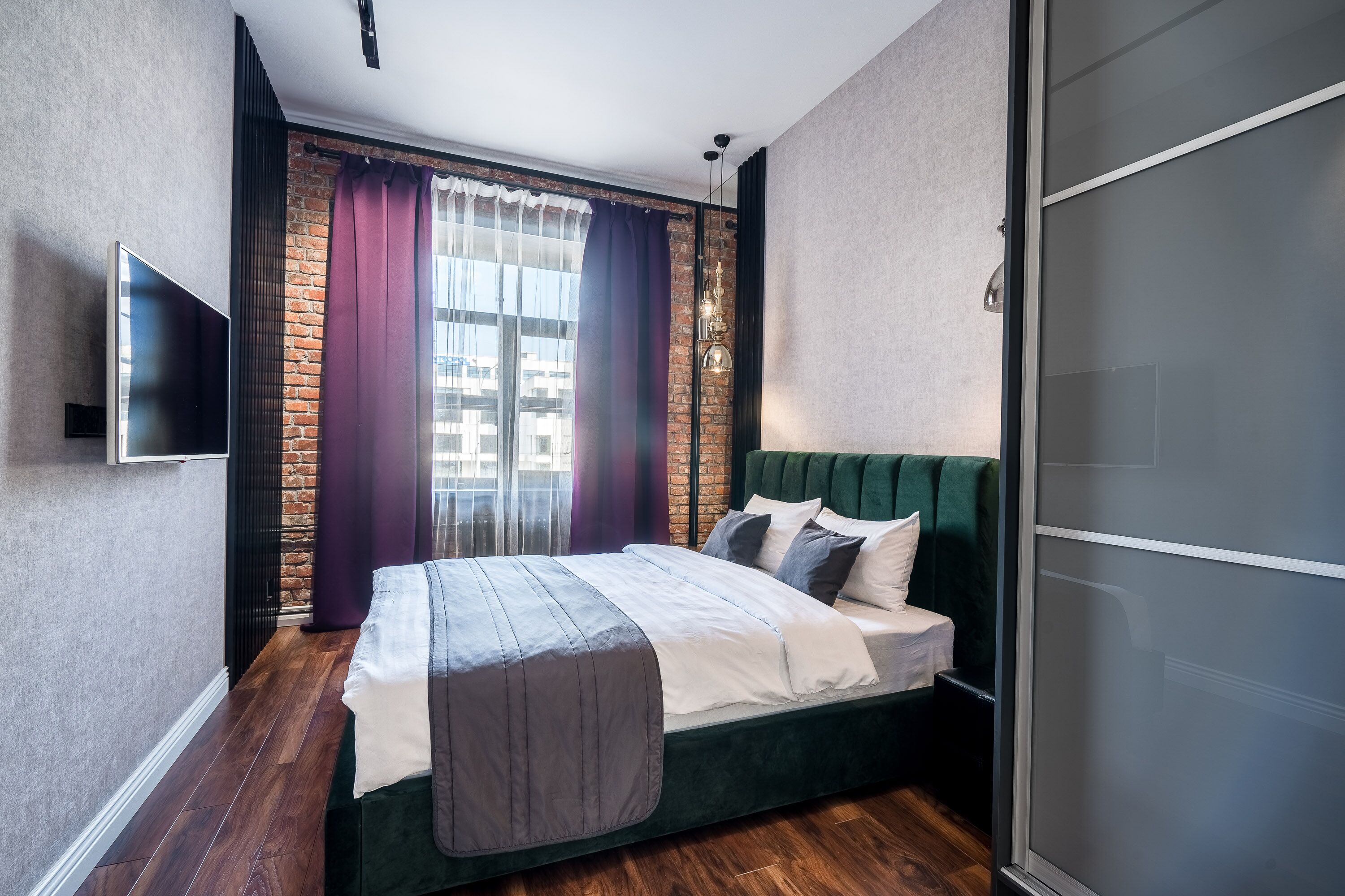 Ein modernes Schlafzimmer mit Backsteinwand, einem grünen Stoffpolster-Kopfstück, einem Flachbildfernseher und einem großen Fenster mit violetten Vorhängen.